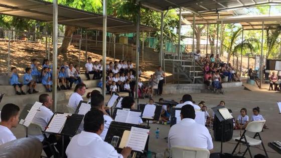 Se observa de espalda a los músicos y frente a ellos niñas y niños sentados en las gradas de un gimnasio y también en el suelo frente a ellos ríen o disfrutan admirados del concierto. Niñas y niños de todas las edades rodean la Banda a los lados del gimnasio. Hermosa luz de día llena de calidez el ambiente del concierto.