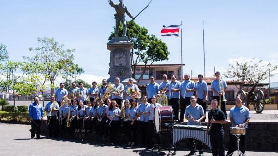 En la foto se aprecia a los músicos de la Banda de Conciertos de Alajuela junto a sus instrumentos, posando en el parque Juan Santa María con la estatua de fondo. Visten con pantalón negro y camisa azul, todos sonríen a la luz de la mañana.