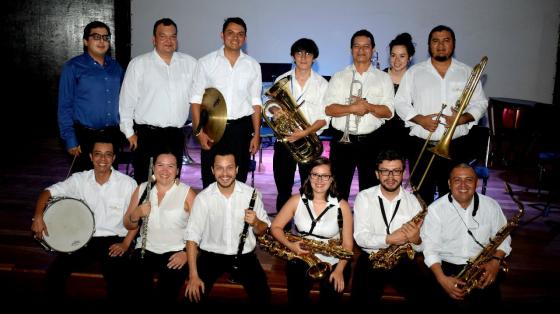 Concierto de Bien Social - Banda de Conciertos de Limón La Banda de Conciertos de Limón posando en un escenario vestidos de blanco y negro sonríen sosteniendo sus instrumentos entre las manos.