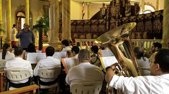 Músicos de la Banda de Conciertos de Alajuela tocando en la catedral de Alajuela. En la foto se aprecia un fondo de color claro dorado con imágenes religiosas alrededor del ensamble y en el centro al director, dirigiendo el concierto.