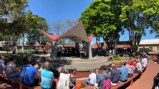 Concierto de celebración de la Esparzanidad - Banda de Conciertos de Puntarenas. La Banda de Conciertos de Puntarenas toca en un kiosco pequeño visto desde lejos. Público de todas las edades rodean bajo la luz de la mañana, el concierto.
