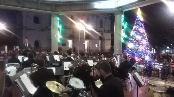 Concierto Popular y Navideño con el Coro del Adulto Mayor UCR - Banda de Conciertos de Heredia Banda de Conciertos de Heredia tocando en el kiosco del parque central de Heredia rodeados de adornos navideños con luces de colores y al fondo un gran árbol navideño lleno de luz junto a la Iglesia La Inmaculada Concepción..