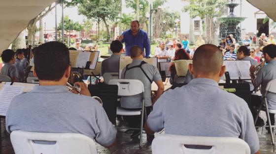 Músicos de la Banda de Conciertos de Alajuela sentados, tocando un concierto en el kiosco del parque central de Alajuela, dirigidos por Do Gabriel Campos. Concierto de día en el que se aprecia al público disfrutando del concierto.