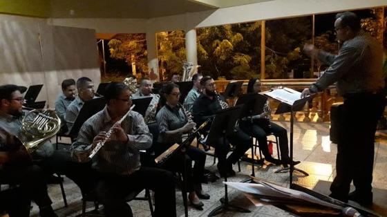 Concierto Especial - Banda de Conciertos de Puntarenas. Músicos tocan sus instrumentos en un salón pequeño. Visten su uniforme de rayas blancas y negras. Sentados con sus instrumentos ejecutan las indicaciones del director.