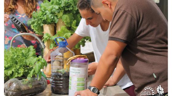 Dos jóvenes realizando compostaje, al frente cuentan con 2 botellas y una de ella dice material compostear, el otro es una botella con pequeñas lechugas