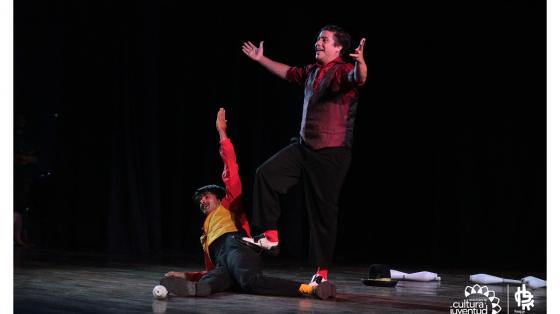 Celebración del Día Mundial del Circo Social Dos chicos haciendo el acto de cierre de una presentación de circo, ambos con colores vistoso y atrás de ellos como unos pinos de boliche
