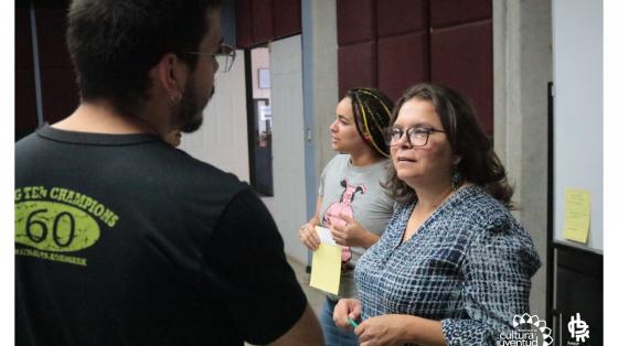 Dos personas conversando sobre un tema en especifico, detrás de ellos una muchacha con una pizarra al lado