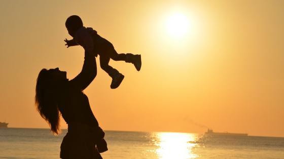 Mujer madre sostiene a un niño entre sus brazos, detrás de ella la luz del atardecer con el mar de fondo.
