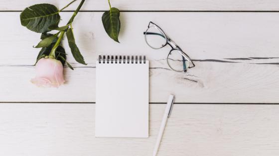 Escritorio con una rosa blanca, una libreta y unos lentes