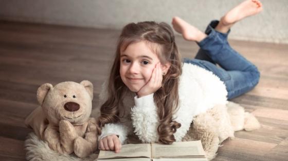 Niña leyendo un libro junto a un oso de peluche