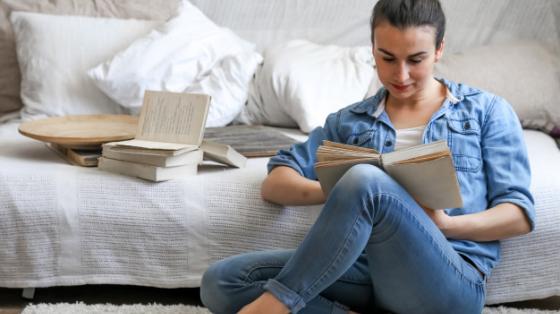 Chica leyendo un libro en el piso y detrás la cama y algunos libros