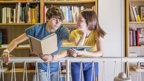 Dos jóvenes hablando en la biblioteca mientras leen.