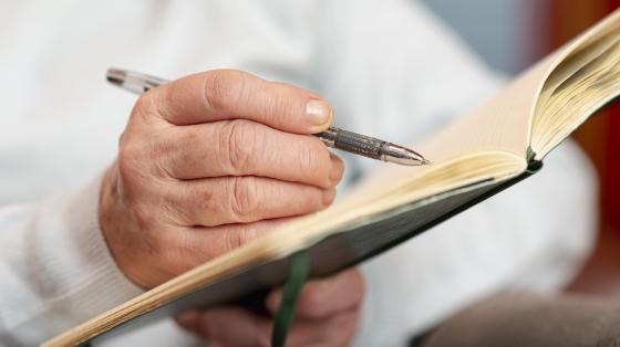 Tertulia, voz y pluma: personas adultas mayores en la escritura Mano de persona adulta mayor escribiendo en un libro.