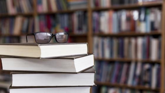 Libros apilados Fotografía con un fondo en segundo plano de estaterías de libros. En el frente de la fotografía se encuentran 4 libros apilados y encima de estos un par de gafas.