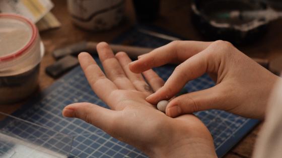 Manualidad Fotografía donde se muestran las manos de una persona realizando una manualidad, en el fondo se observa un espacio con pinceles, papeles y artículos de manualidades.