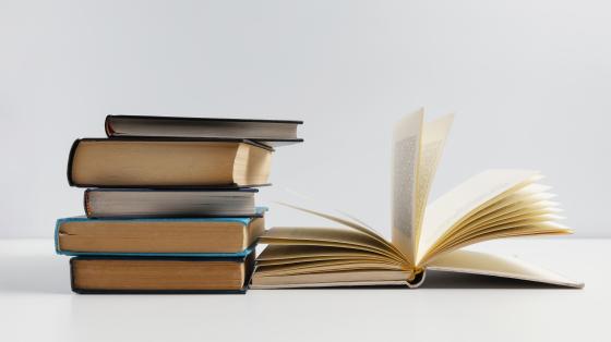 Fotografía con fondo gris, se observan 5 libros cerrados apilados unos sobre otros, a su lado hay un libro abierto a la mitad.