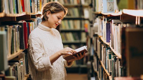 Fotografía de una mujer leyendo un libro en medio de dos estanterías de libros.