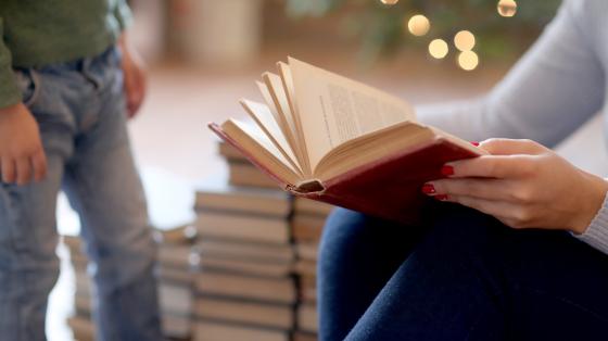 Cuentacuentos para los más pequeñitos Persona sosteniendo un libro abierto y a la izquierda un niño. Fondo navideño.