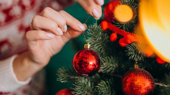 Manos decorando un árbol de navidad con ornamentos circulares rojos. 