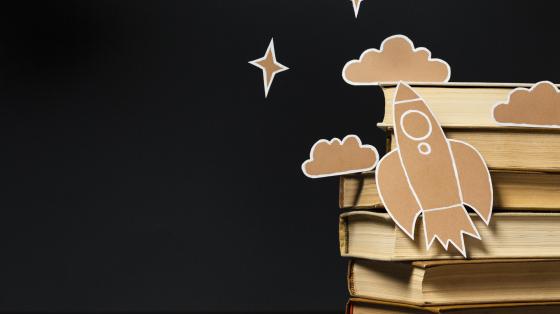Imagen con fondo negro al lado derecho unos libros apilados y sobre los libros unas figuras en cartón de un cohete, unas nubes y estrellas