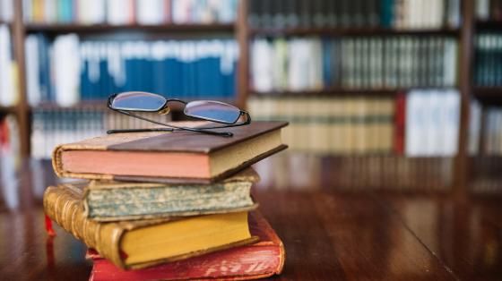 Conferencia "El paraíso en llamas: 75 años de la Guerra Civil de 1948 en Costa Rica" Fondo de una biblioteca, sobre una mesa de madera una torre de libros y encima unos anteojos.