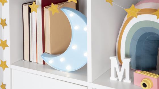 Imagen de un estante con repisas color blanco, decorado con estrellas doradas, en un estante unos libros junto a una figura de luna con luz, en el otro estante se observa una letra M, una cámara de juguete y un almohadón en forma de arcoiris.