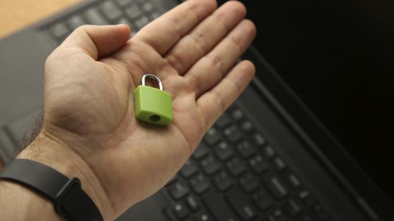 Charla "Ciudadanía digital" En el fono una computadora portátil sobre una mesa de madera, y encima una manos sosteniendo un candado.