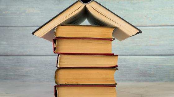 Sobre fondo de madera, se encuentra una torre de libros y encima un libro abierto.
