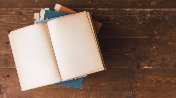 Sobre fondo de madera, tres libros formando una torre y encima un libro abierto. 