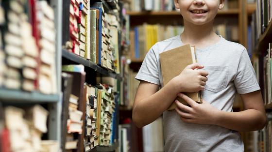 Arcoíris de Lectura Un niño sosteniendo un libro en medio de una colección de libros.