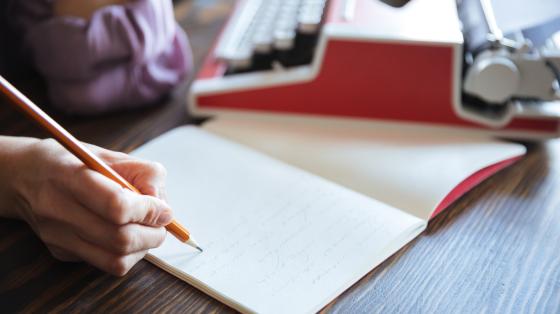 Sobre una mesa de madera, una persona escribiendo en un libro y a la par una máquina de escribir.