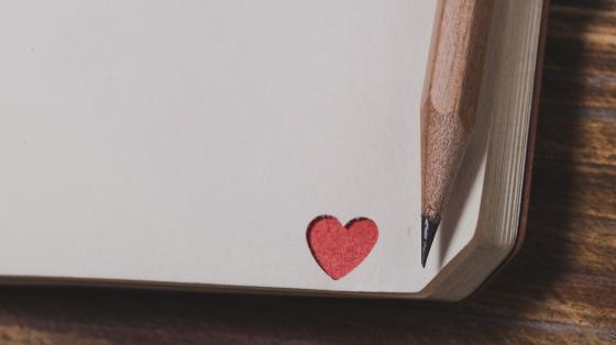 Celebración del Día Nacional de la Poesía Fondo de madera. Esquina de un libro con un corazón rojo y un lápiz de grafito a la derecha.