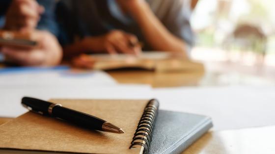 Personas de fondo revisando un libro y en primer plano unos libros y una plumilla