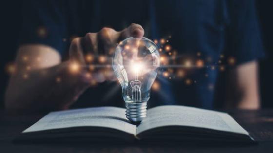 Exposición sobre Nikola Tesla Un libro abierto y un bombillo sobre el iluminado y una persona detrás tocándolo con el dedo índice