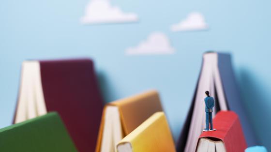 De fondo un cielo con unas nubes, varios libros entreabiertos con el lomo hacia arriba y una figura de una persona sobre uno de los libros