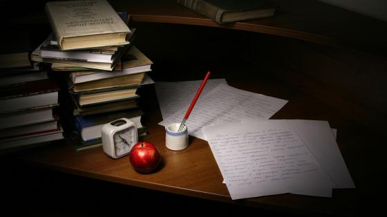 Taller "Incorporación del lenguaje inclusivo en el texto literario" Sobre un escritorio varios libros apilados, unas hojas escritas a manos, una pluma para escribir, un reloj y una manzana
