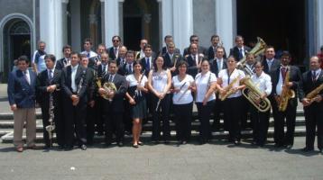 Músicos y músicas del ensamble de la Banda de Cartago frente a una iglesia grande y blanca. Todo el grupo sonríe sosteniendo sus instrumentos.
