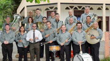 El ensamble musical completo junto a sus instrumentos posa de  pie frente a la cámara, detrás de ellos en ambiente exterior hay una casa blanca antigua de fondo. Todas y todos lucen muy sonrientes.