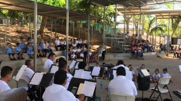 Se observa de espalda a los músicos y frente a ellos niñas y niños sentados en las gradas de un gimnasio y también en el suelo frente a ellos ríen o disfrutan admirados del concierto. Niñas y niños de todas las edades rodean la Banda a los lados del gimnasio. Hermosa luz de día llena de calidez el ambiente del concierto.