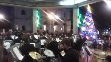 Banda de Conciertos de Heredia tocando en el kiosco del parque central de Heredia rodeados de adornos navideños con luces de colores y al fondo un gran árbol navideño lleno de luz junto a la Iglesia La Inmaculada Concepción..
