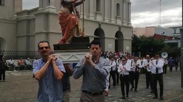 En el parque de Heredia unos hombres llevan en sus hombros un anda religiosa, atrás acompañan con música el ensamble de la Banda de Conciertos de Heredia recorriendo a pie mientras tocan para acompañar a la procesión.