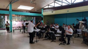 Músicos de la Banda de Conciertos tocando dentro de un salón tipo galerón con fondo azul. Se aprecia un lugar amplio lleno de luz mientras ellos realizan su concierto.