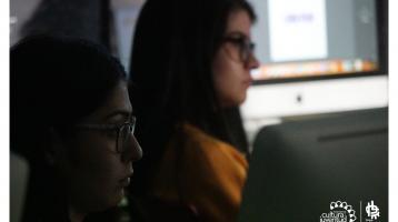 Dos jóvenes prestando atención a una clase, detrás de ellas varias computadoras