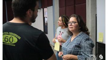 Dos personas conversando sobre un tema en especifico, detrás de ellos una muchacha con una pizarra al lado