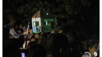 Grupo de personas en el desfile de faroles, sobresale un farol en forma de casita.