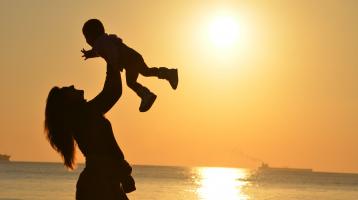 Mujer madre sostiene a un niño entre sus brazos, detrás de ella la luz del atardecer con el mar de fondo.