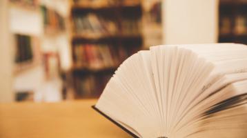 Libro abierto y en el fondo unos estantes de biblioteca con libros