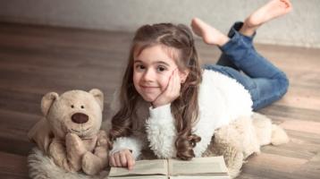 Niña leyendo un libro junto a un oso de peluche