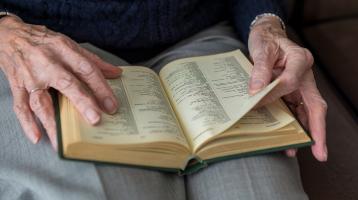 Fotografía de una persona adulta mayor leyendo un libro. En la fotografía solo se hace un plano del regazo de la persona donde se ve el libro y sus manos sosteniéndolo.  
