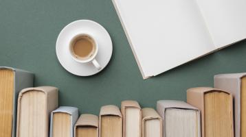 Fotografía en la cual en la esquina superior derecha se encuentra un libro abierto a la mitad, a su lado una taza. En la parte inferior de la fotografía se encuentran diez libros acomodados de lado uno con otros